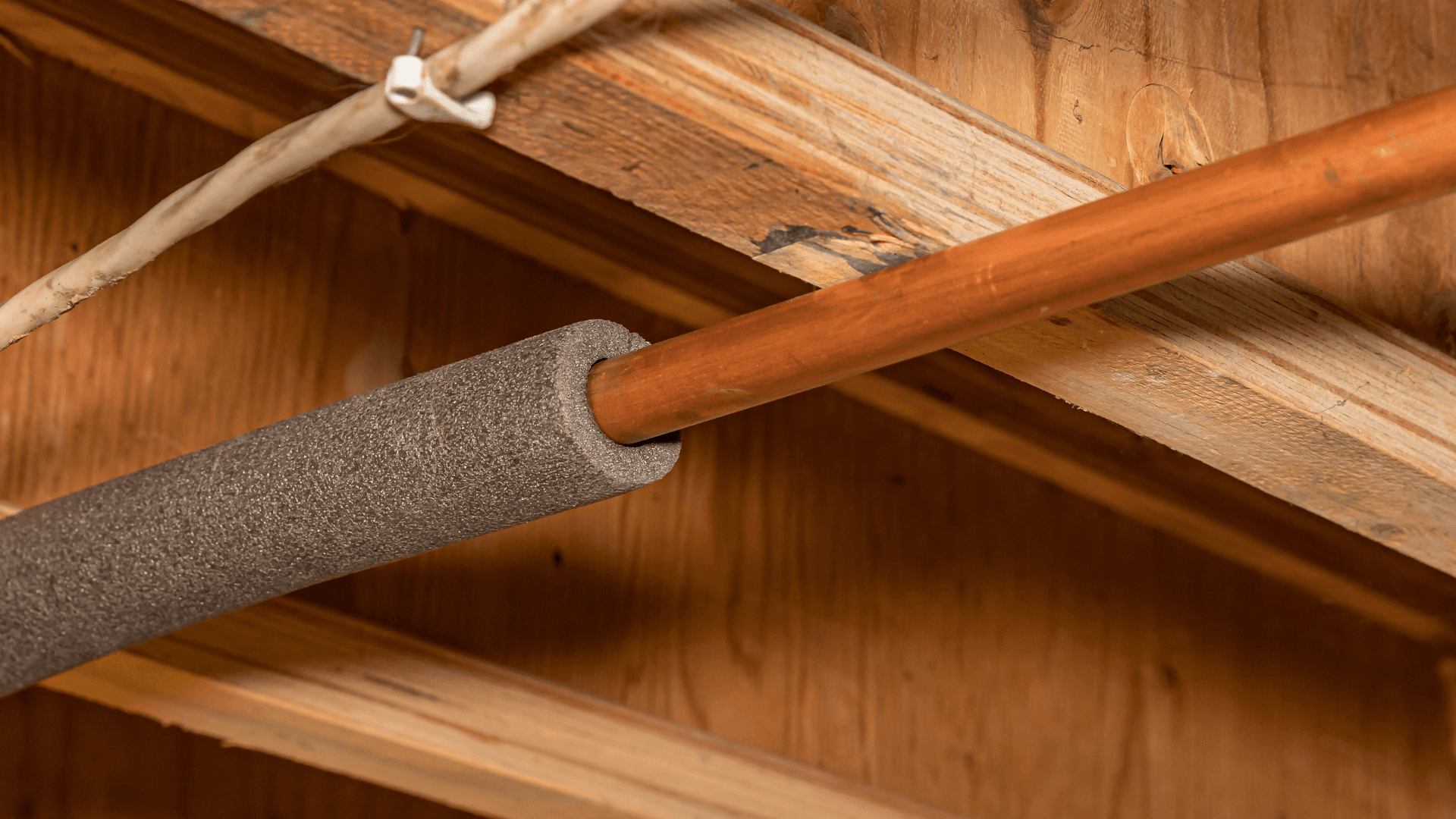 Close-up of a copper pipe partly covered by gray foam insulation beneath wooden rafters, with a white cable secured overhead; rustic utility scene.