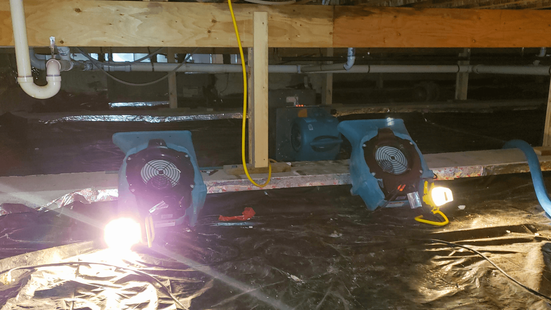 Dim crawlspace with blue ventilation fans, bright work lights, plastic sheeting, exposed pipes, beams, and a yellow electrical cord.