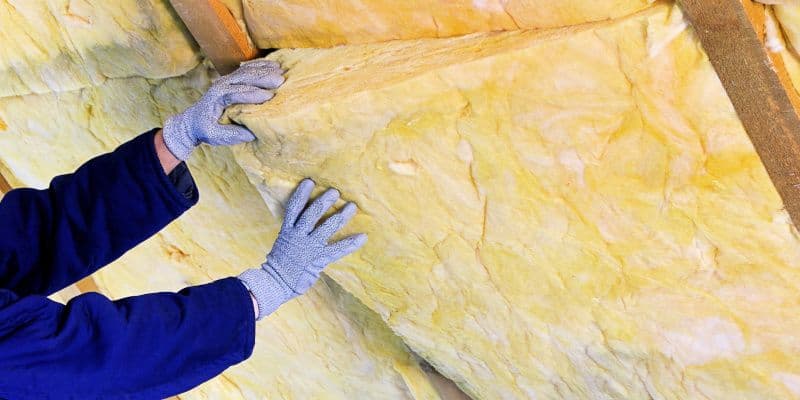 Gloved worker installs yellow fiberglass insulation between wooden attic beams, pressing the material into place for home energy efficiency.