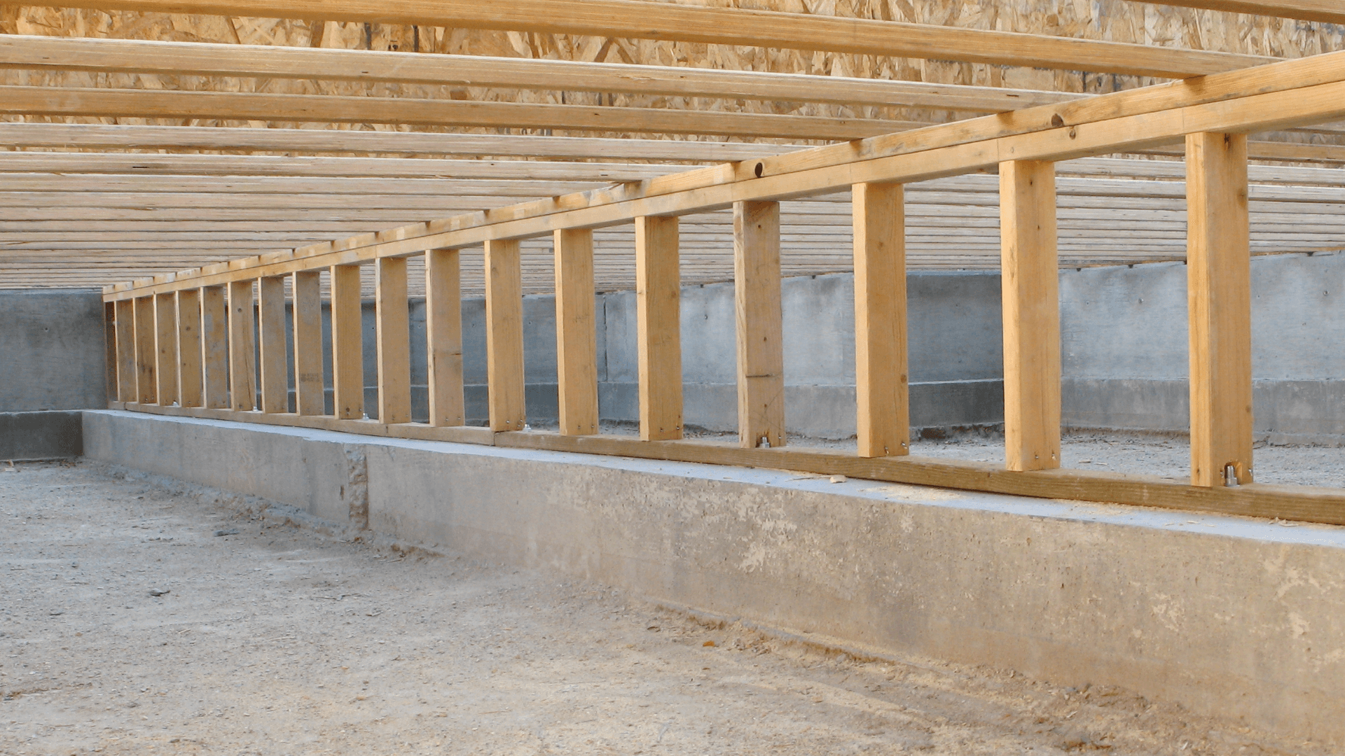 Unfinished wooden building frame with evenly spaced posts and roof beams on a concrete foundation, viewed in perspective; neutral construction scene.