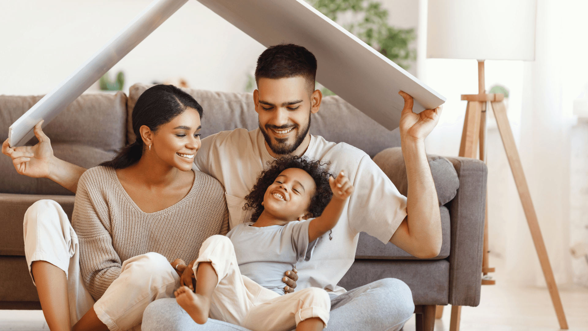 Smiling parents cuddle their laughing child beneath a couch-cushion fort, creating a warm, playful family moment in a bright living room.