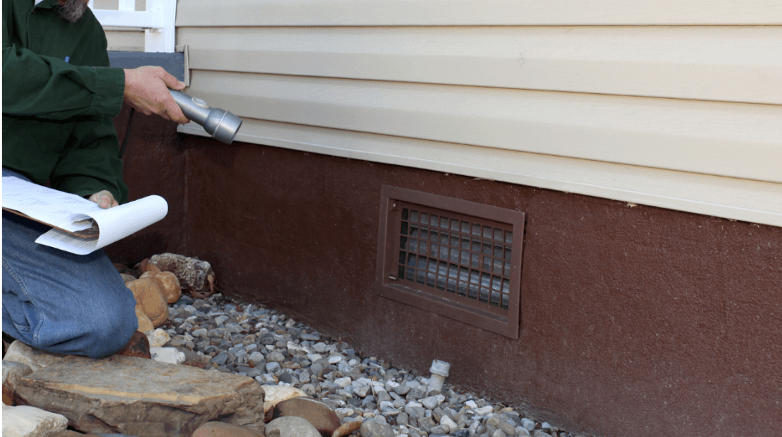 Home inspector shines a flashlight at a barred foundation vent beneath beige siding, holding notes beside a gravel bed during a routine inspection.