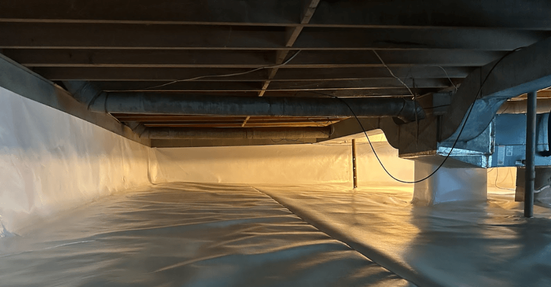 Low crawlspace beneath a building, lined with plastic vapor barriers and crossed by dark beams, ducts, and pipes in warm sunlight.